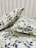 Two pillows are placed on a bed with a matching floral-patterned fabric. The fabric features a botanical design with leaves and branches in shades of gray against a light cream background. The headboard is tufted and covered in a soft, light-colored material.