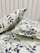 Two pillows are placed on a bed with a matching floral-patterned fabric. The fabric features a botanical design with leaves and branches in shades of gray against a light cream background. The headboard is tufted and covered in a soft, light-colored material.