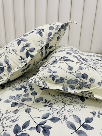 Two pillows are placed on a bed with a matching floral-patterned fabric. The fabric features a botanical design with leaves and branches in shades of gray against a light cream background. The headboard is tufted and covered in a soft, light-colored material.