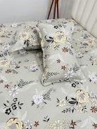 A bed neatly covered with a floral-patterned bedsheet and matching pillowcases. The patterns feature various types of flowers in shades of yellow, brown, black, and white, set against a light gray background. A wooden tripod stand is positioned near the bed, partially visible.