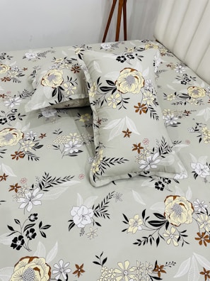 A bed neatly covered with a floral-patterned bedsheet and matching pillowcases. The patterns feature various types of flowers in shades of yellow, brown, black, and white, set against a light gray background. A wooden tripod stand is positioned near the bed, partially visible.