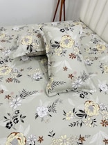 A bed neatly covered with a floral-patterned bedsheet and matching pillowcases. The patterns feature various types of flowers in shades of yellow, brown, black, and white, set against a light gray background. A wooden tripod stand is positioned near the bed, partially visible.