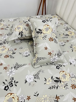 A bed neatly covered with a floral-patterned bedsheet and matching pillowcases. The patterns feature various types of flowers in shades of yellow, brown, black, and white, set against a light gray background. A wooden tripod stand is positioned near the bed, partially visible.