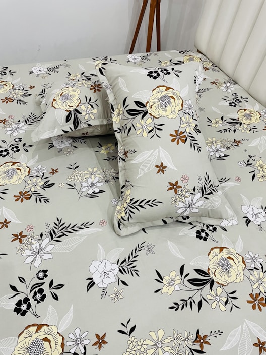 A bed neatly covered with a floral-patterned bedsheet and matching pillowcases. The patterns feature various types of flowers in shades of yellow, brown, black, and white, set against a light gray background. A wooden tripod stand is positioned near the bed, partially visible.