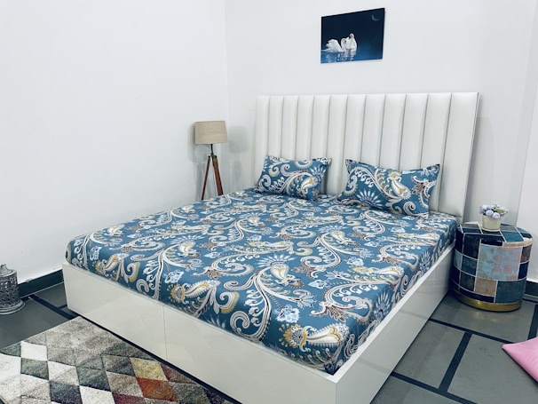 A neatly made bed with a blue and white floral pattern on the bedspread is situated in a modern bedroom. The headboard is white and vertically paneled. To the side, there is a wooden floor lamp and a small round side table with a colorful mosaic design. A small potted plant with purple flowers is placed on the table. A decorative carpet with a geometric pattern lies on the floor, and a framed picture of swans on water hangs on the wall above the bed.