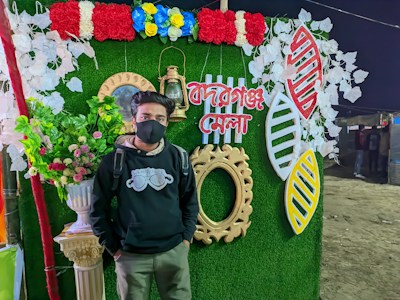 A person wearing a black hoodie and face mask stands in front of a decorative backdrop featuring vibrant red, white, and blue artificial flowers, leaves, and ornate items. The backdrop includes text in a script and two large cutouts resembling artistic shapes, all set against a green background. A decorative lantern and a circular frame further enhance the setup, and there's a clay column with a bouquet of mixed flowers.
