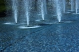 Several water fountains are arranged in a row, jetting streams of water high into the air above a calm pool with a textured surface. The water appears clear, and the overall atmosphere is serene and peaceful. Surrounding the fountains, there is a grassy area and some trees that provide a natural backdrop.