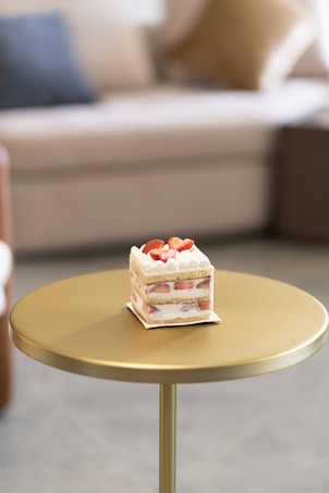 A slice of cake with layers of cream and strawberries is placed on a small, round, golden table. The background includes a blurred image of a couch with cushions, creating a cozy and inviting atmosphere.