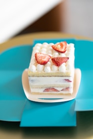 A square layered cake with white cream and strawberries on top, presented on a light pink plate. The cake includes layers of white cream and sponge, with visible strawberry slices inside. It is placed on a teal green surface that contrasts with the cake.