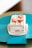 A square layered cake with white cream and strawberries on top, presented on a light pink plate. The cake includes layers of white cream and sponge, with visible strawberry slices inside. It is placed on a teal green surface that contrasts with the cake.