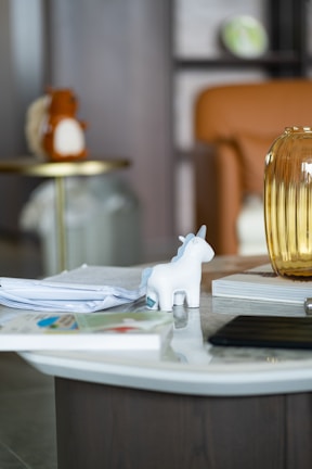 A white unicorn figurine with a blue mane and horn is placed on a round coffee table. The table also has a stack of papers, a book, and a transparent amber glass vase or jar. In the background, there is a blurred orange chair and a round side table holding a soft toy or cushion resembling an animal. The setting appears to be a cozy indoor space, possibly a living room.