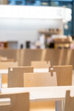 A bright, modern cafeteria setting featuring rows of light wooden chairs and tables. The background is blurred with hints of a counter or bar area, giving a clean and minimalistic feel.