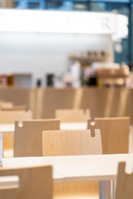 A bright, modern cafeteria setting featuring rows of light wooden chairs and tables. The background is blurred with hints of a counter or bar area, giving a clean and minimalistic feel.