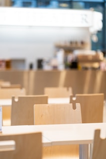 A bright, modern cafeteria setting featuring rows of light wooden chairs and tables. The background is blurred with hints of a counter or bar area, giving a clean and minimalistic feel.