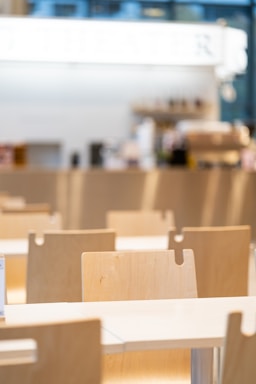 A bright, modern cafeteria setting featuring rows of light wooden chairs and tables. The background is blurred with hints of a counter or bar area, giving a clean and minimalistic feel.
