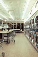 A laundromat interior featuring a row of industrial washing machines on the right side and several laundry carts lined up on the left. The ceiling is adorned with white panels and fluorescent lights. Tables for folding laundry are positioned between the carts. The floor has a speckled pattern, and the overall atmosphere is clean and orderly.