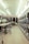 A laundromat interior featuring a row of industrial washing machines on the right side and several laundry carts lined up on the left. The ceiling is adorned with white panels and fluorescent lights. Tables for folding laundry are positioned between the carts. The floor has a speckled pattern, and the overall atmosphere is clean and orderly.