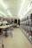 A laundromat interior featuring a row of industrial washing machines on the right side and several laundry carts lined up on the left. The ceiling is adorned with white panels and fluorescent lights. Tables for folding laundry are positioned between the carts. The floor has a speckled pattern, and the overall atmosphere is clean and orderly.