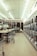 A laundromat interior featuring a row of industrial washing machines on the right side and several laundry carts lined up on the left. The ceiling is adorned with white panels and fluorescent lights. Tables for folding laundry are positioned between the carts. The floor has a speckled pattern, and the overall atmosphere is clean and orderly.