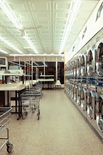 A laundromat interior featuring a row of industrial washing machines on the right side and several laundry carts lined up on the left. The ceiling is adorned with white panels and fluorescent lights. Tables for folding laundry are positioned between the carts. The floor has a speckled pattern, and the overall atmosphere is clean and orderly.