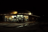 A laundromat illuminated at night, with bright fluorescent lights inside revealing rows of washing machines and chairs. The parking lot is dark and the pavement is wet, possibly from rain.