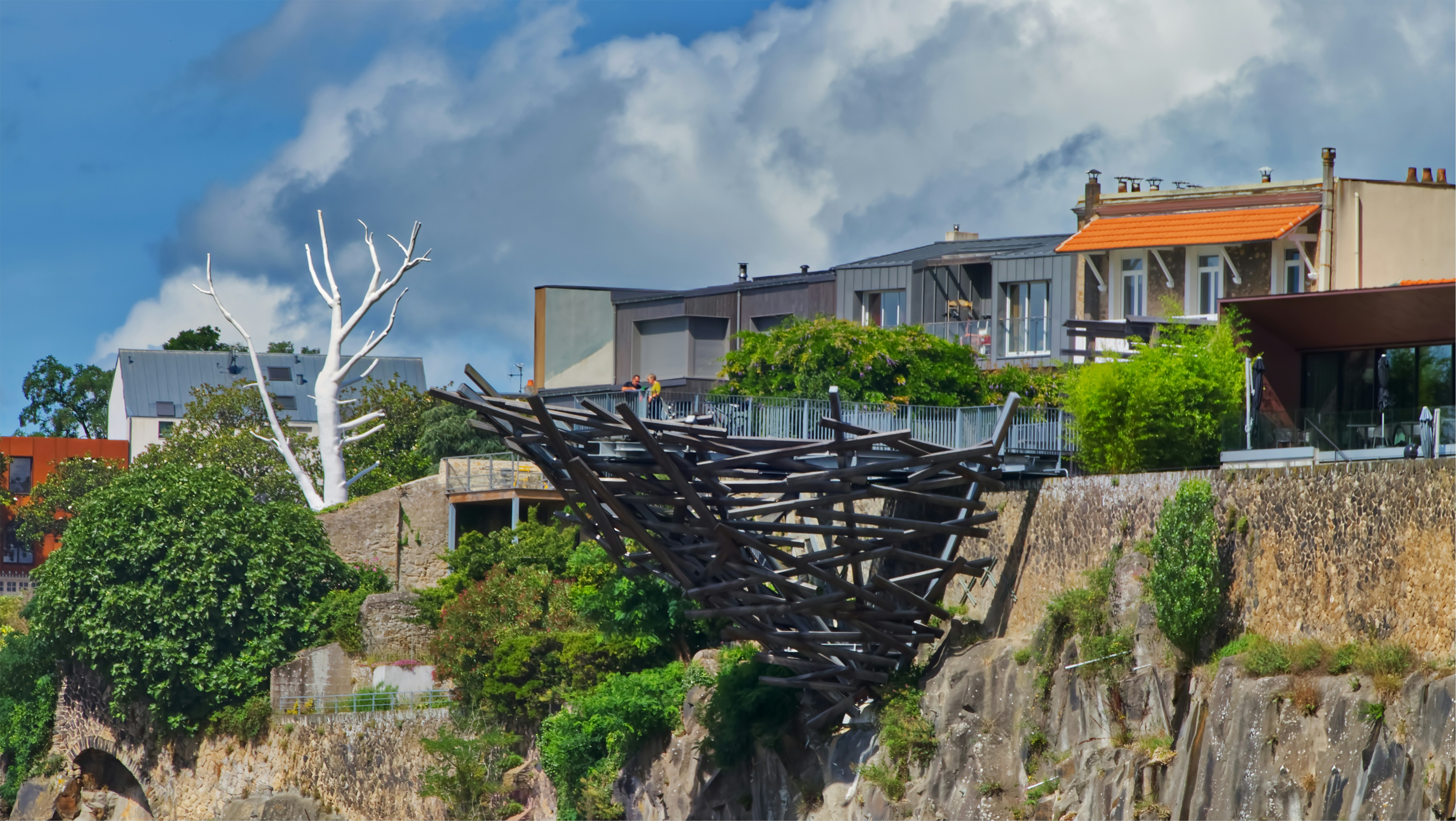 a sculpture on the side of a cliff with houses in the background, Vue sur le Nid de Jules Verne et sur l’arbre blanc de Númenor sur le Quai Marquis d’Aiguillon à Nantes. Jules Verne est natif de Nantes et l’arbre blanc représente un symbole de la Saga du Seigneurs des Anneaux.