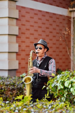 A person wearing sunglasses and a hat is playing a saxophone near a brick building. Surrounding them are green shrubs, adding a natural element to the urban setting.