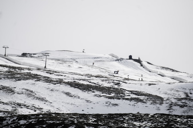 A snow-covered mountainous landscape with visible ski slopes and ski lifts. The terrain is marked by patches of exposed earth and stretches of snow, with outlines of fencing or barriers running along the slope. There are a few small structures on the hill, potentially ski huts or lift stations.