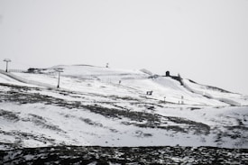 A snow-covered mountainous landscape with visible ski slopes and ski lifts. The terrain is marked by patches of exposed earth and stretches of snow, with outlines of fencing or barriers running along the slope. There are a few small structures on the hill, potentially ski huts or lift stations.