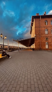 A railway station platform featuring a large, historic brick building with arched windows. The sky is a striking mix of deep blue and grey clouds, suggesting a dramatic atmosphere. A person sits on a yellow bench on the platform, adding a sense of solitude or contemplation. The platform is lined with street lamps, and a train is visible in the background.