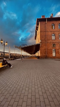 A railway station platform featuring a large, historic brick building with arched windows. The sky is a striking mix of deep blue and grey clouds, suggesting a dramatic atmosphere. A person sits on a yellow bench on the platform, adding a sense of solitude or contemplation. The platform is lined with street lamps, and a train is visible in the background.