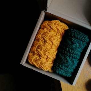 A cardboard box containing two knitted items, one in vibrant orange and the other in deep green, placed on a wooden surface. The lighting casts a shadow on the left, highlighting the texture of the yarn.