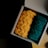 A cardboard box containing two knitted items, one in vibrant orange and the other in deep green, placed on a wooden surface. The lighting casts a shadow on the left, highlighting the texture of the yarn.