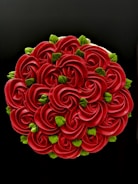 An intricately designed cake with a floral pattern, featuring a series of red rose-like shapes made from frosting, interspersed with small, bright green leaf decorations. The black background enhances the vividness of the colors.