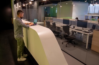A man stands at a curved white counter using a laptop, with a modern office visible behind a glass wall. The office has several workstations with large monitors, chairs, and another person focused on their computer. The lighting is soft, creating a calm atmosphere.