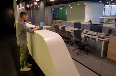 A man stands at a curved white counter using a laptop, with a modern office visible behind a glass wall. The office has several workstations with large monitors, chairs, and another person focused on their computer. The lighting is soft, creating a calm atmosphere.
