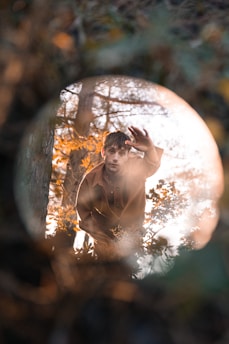 A person is reaching out towards the camera with a hand raised. The setting appears to be outdoors with trees and foliage, creating a natural and serene environment. Warm sunlight filters through the branches, casting a soft glow on the scene.