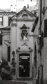 A narrow, quaint alleyway leads to a ceramic-style building featuring an arched doorway with ornate detailing above it. The door itself is dark, with a sign that reads 'Ristorante Pizzeria'. The surrounding architecture is traditional, with rustic charm. Two decorative street lamps are affixed to the facade and walls. There are small potted plants and greenery visible, adding a touch of nature to the otherwise stone-and-brick environment.