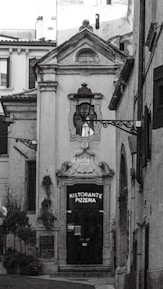 A narrow, quaint alleyway leads to a ceramic-style building featuring an arched doorway with ornate detailing above it. The door itself is dark, with a sign that reads 'Ristorante Pizzeria'. The surrounding architecture is traditional, with rustic charm. Two decorative street lamps are affixed to the facade and walls. There are small potted plants and greenery visible, adding a touch of nature to the otherwise stone-and-brick environment.