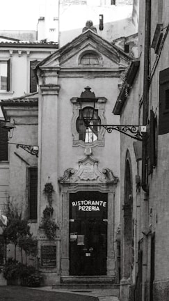 A narrow, quaint alleyway leads to a ceramic-style building featuring an arched doorway with ornate detailing above it. The door itself is dark, with a sign that reads 'Ristorante Pizzeria'. The surrounding architecture is traditional, with rustic charm. Two decorative street lamps are affixed to the facade and walls. There are small potted plants and greenery visible, adding a touch of nature to the otherwise stone-and-brick environment.