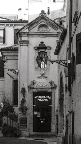 A narrow, quaint alleyway leads to a ceramic-style building featuring an arched doorway with ornate detailing above it. The door itself is dark, with a sign that reads 'Ristorante Pizzeria'. The surrounding architecture is traditional, with rustic charm. Two decorative street lamps are affixed to the facade and walls. There are small potted plants and greenery visible, adding a touch of nature to the otherwise stone-and-brick environment.