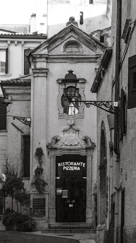 A narrow, quaint alleyway leads to a ceramic-style building featuring an arched doorway with ornate detailing above it. The door itself is dark, with a sign that reads 'Ristorante Pizzeria'. The surrounding architecture is traditional, with rustic charm. Two decorative street lamps are affixed to the facade and walls. There are small potted plants and greenery visible, adding a touch of nature to the otherwise stone-and-brick environment.