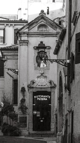 A narrow, quaint alleyway leads to a ceramic-style building featuring an arched doorway with ornate detailing above it. The door itself is dark, with a sign that reads 'Ristorante Pizzeria'. The surrounding architecture is traditional, with rustic charm. Two decorative street lamps are affixed to the facade and walls. There are small potted plants and greenery visible, adding a touch of nature to the otherwise stone-and-brick environment.