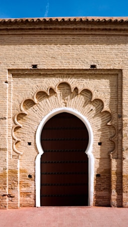 A large, intricately designed Moorish arch made of brick, featuring a decorative pattern that frames a dark wooden door. The arch has an elegant scalloped design and is set against a sunlit brick wall with a terracotta tiled roof above.