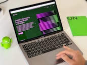 A laptop displaying a webpage about ChatGPT with a green and black design sits open on a desk. Next to the laptop is a small green Android figurine and a green notebook with 'ON' written on it. A hand is visible on the right side, interacting with the laptop's trackpad.