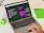 A laptop displaying a webpage about ChatGPT with a green and black design sits open on a desk. Next to the laptop is a small green Android figurine and a green notebook with 'ON' written on it. A hand is visible on the right side, interacting with the laptop's trackpad.