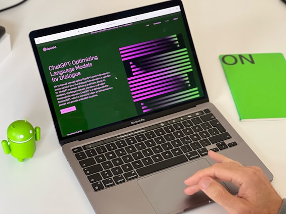 A laptop displaying a webpage about ChatGPT with a green and black design sits open on a desk. Next to the laptop is a small green Android figurine and a green notebook with 'ON' written on it. A hand is visible on the right side, interacting with the laptop's trackpad.