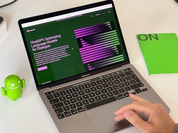 A laptop displaying a webpage about ChatGPT with a green and black design sits open on a desk. Next to the laptop is a small green Android figurine and a green notebook with 'ON' written on it. A hand is visible on the right side, interacting with the laptop's trackpad.
