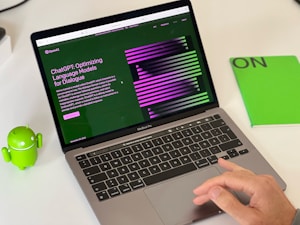 A laptop displaying a webpage about ChatGPT with a green and black design sits open on a desk. Next to the laptop is a small green Android figurine and a green notebook with 'ON' written on it. A hand is visible on the right side, interacting with the laptop's trackpad.