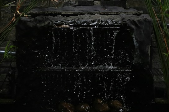 A dark water fountain with cascading water flowing down a rugged, stone-like structure. The backdrop consists of a textured stone wall with some greenery, including tall leaves and plants framing the fountain.